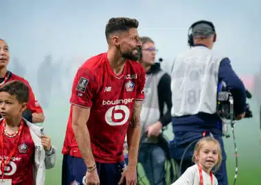 Bearded soccer player in a red jersey bends slightly forward on a field, surrounded by children and a filming crew in the background.