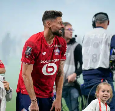 Bearded soccer player in a red jersey bends slightly forward on a field, surrounded by children and a filming crew in the background.