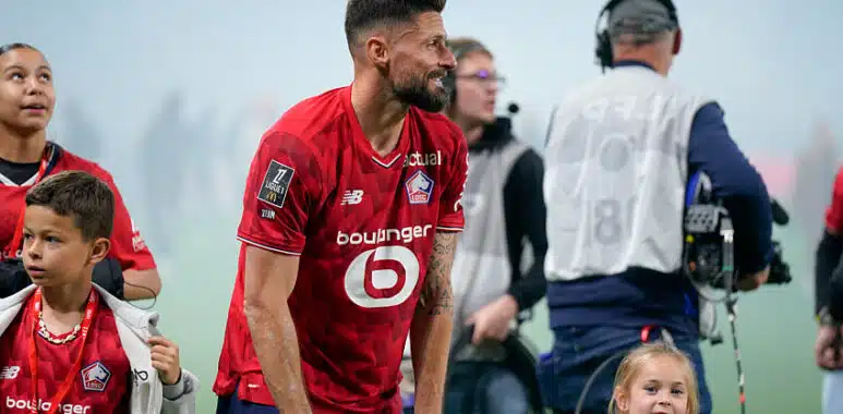 Giroud | AllezPaillade.com Bearded soccer player in a red jersey bends slightly forward on a field, surrounded by children and a filming crew in the background.