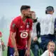 Bearded soccer player in a red jersey bends slightly forward on a field, surrounded by children and a filming crew in the background.
