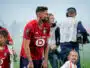 Bearded soccer player in a red jersey bends slightly forward on a field, surrounded by children and a filming crew in the background.
