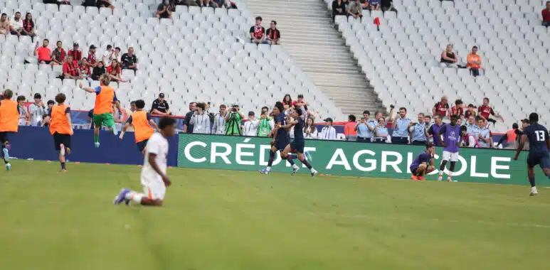 Photo by Icon Sport - Photo by Icon Sport | AllezPaillade.com Soccer players in orange training vests run on the field as spectators watch from white seats in a stadium.