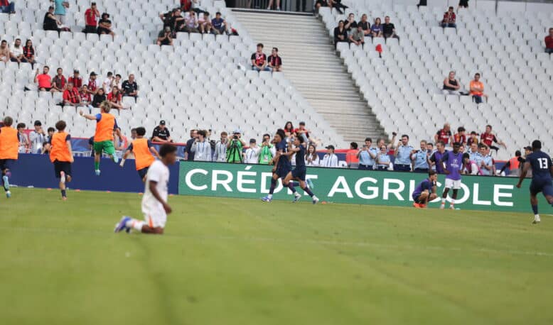 Soccer players in orange training vests run on the field as spectators watch from white seats in a stadium.