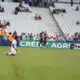 Soccer players in orange training vests run on the field as spectators watch from white seats in a stadium.