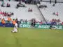 Soccer players in orange training vests run on the field as spectators watch from white seats in a stadium.