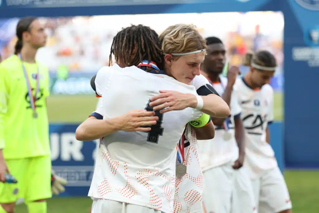 Two athletes in white uniforms hug in celebration on a sports field, medal visible at the neck, blurred crowd in the background.