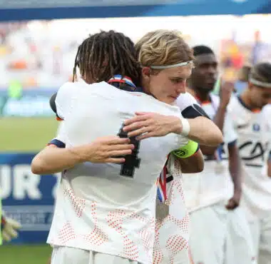 Two athletes in white uniforms hug in celebration on a sports field, medal visible at the neck, blurred crowd in the background.