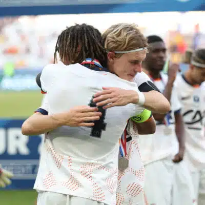 Two athletes in white uniforms hug in celebration on a sports field, medal visible at the neck, blurred crowd in the background.