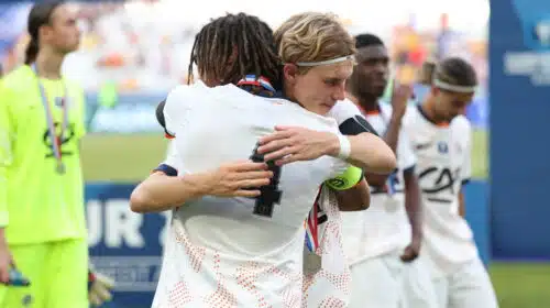 Two athletes in white uniforms hug in celebration on a sports field, medal visible at the neck, blurred crowd in the background.