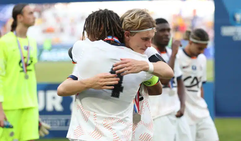 Two athletes in white uniforms hug in celebration on a sports field, medal visible at the neck, blurred crowd in the background.