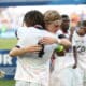 Two athletes in white uniforms hug in celebration on a sports field, medal visible at the neck, blurred crowd in the background.