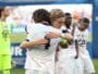 Two athletes in white uniforms hug in celebration on a sports field, medal visible at the neck, blurred crowd in the background.