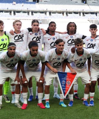 Eleven soccer players pose for a team photo on the field; the goalkeeper wears neon green, others in white kits with red accents, stadium seats in the background.
