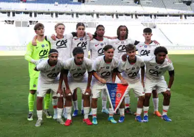 Eleven soccer players pose for a team photo on the field; the goalkeeper wears neon green, others in white kits with red accents, stadium seats in the background.