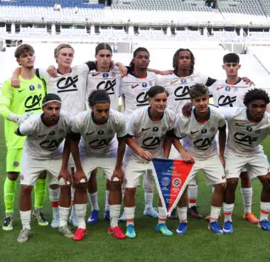 Eleven soccer players pose for a team photo on the field; the goalkeeper wears neon green, others in white kits with red accents, stadium seats in the background.