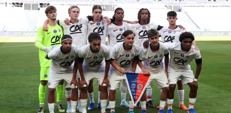 Paris Saint-Germain v Montpellier HSC - Gambardella Credit Agricole Cup Final 2026 | AllezPaillade.com Eleven soccer players pose for a team photo on the field; the goalkeeper wears neon green, others in white kits with red accents, stadium seats in the background.