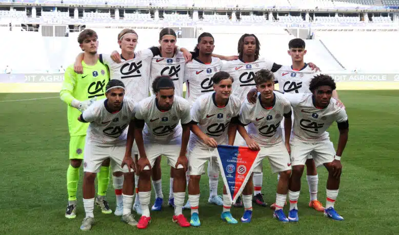 Eleven soccer players pose for a team photo on the field; the goalkeeper wears neon green, others in white kits with red accents, stadium seats in the background.