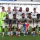 Eleven soccer players pose for a team photo on the field; the goalkeeper wears neon green, others in white kits with red accents, stadium seats in the background.