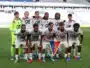 Eleven soccer players pose for a team photo on the field; the goalkeeper wears neon green, others in white kits with red accents, stadium seats in the background.