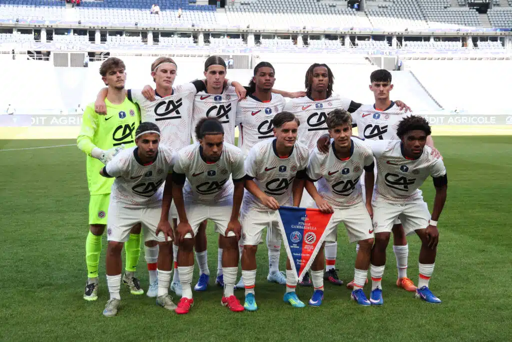 Soccer team photo on the pitch: players in white jerseys with sponsor logos, goalkeeper in neon green, arms around each other in front of empty stadium stands.
