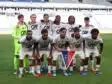 Soccer team photo on the pitch: players in white jerseys with sponsor logos, goalkeeper in neon green, arms around each other in front of empty stadium stands.