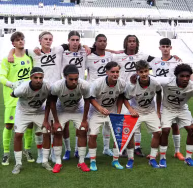 Soccer team photo on the pitch: players in white jerseys with sponsor logos, goalkeeper in neon green, arms around each other in front of empty stadium stands.