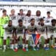 Soccer team photo on the pitch: players in white jerseys with sponsor logos, goalkeeper in neon green, arms around each other in front of empty stadium stands.