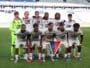 Soccer team photo on the pitch: players in white jerseys with sponsor logos, goalkeeper in neon green, arms around each other in front of empty stadium stands.
