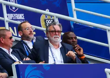 Group of men in suits seated in a stadium VIP box with blue banners behind them, watching the event.
