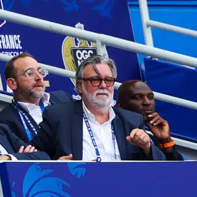 Group of men in suits seated in a stadium VIP box with blue banners behind them, watching the event.