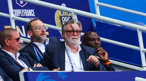 Group of men in suits seated in a stadium VIP box with blue banners behind them, watching the event.