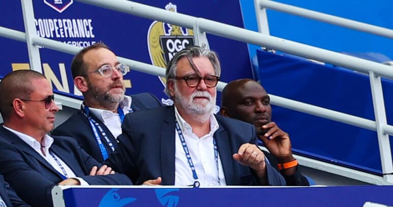 Group of men in suits seated in a stadium VIP box with blue banners behind them, watching the event.