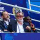 Group of men in suits seated in a stadium VIP box with blue banners behind them, watching the event.