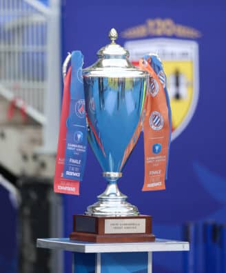 Shiny silver trophy with blue and orange ribbons on a wooden pedestal, set against a blue backdrop (awards scene).