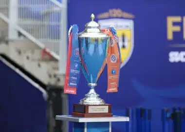 Shiny silver trophy with blue and orange ribbons on a wooden pedestal, set against a blue backdrop (awards scene).