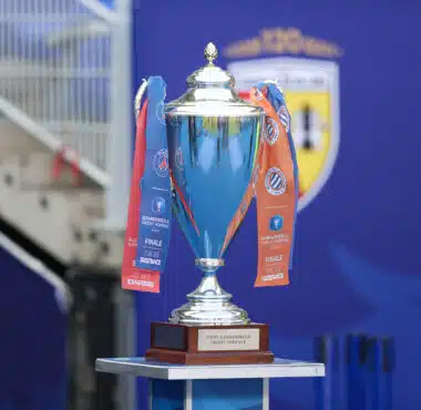 Shiny silver trophy with blue and orange ribbons on a wooden pedestal, set against a blue backdrop (awards scene).