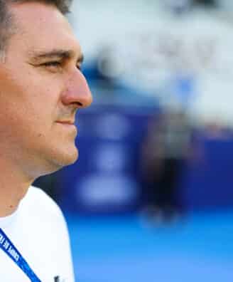 Man in a white shirt shown in profile, wearing a blue lanyard at a sports arena or venue.
