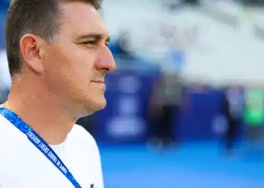Man in a white shirt shown in profile, wearing a blue lanyard at a sports arena or venue.