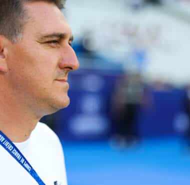 Man in a white shirt shown in profile, wearing a blue lanyard at a sports arena or venue.