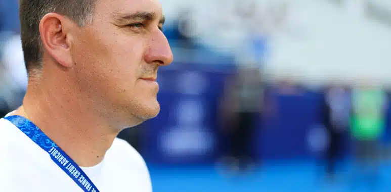 Man in a white shirt shown in profile, wearing a blue lanyard at a sports arena or venue.