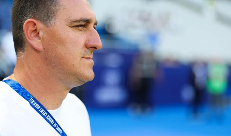 Man in a white shirt shown in profile, wearing a blue lanyard at a sports arena or venue.