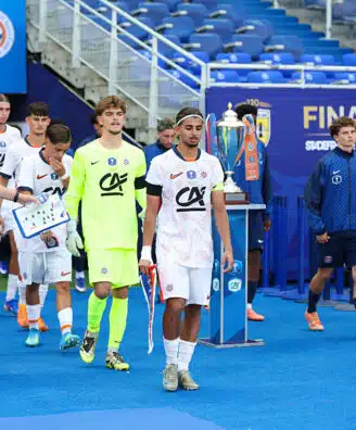 Soccer players in white uniforms walk on a blue field toward a trophy display during a final ceremony, with a neon-clad goalkeeper and staff nearby.