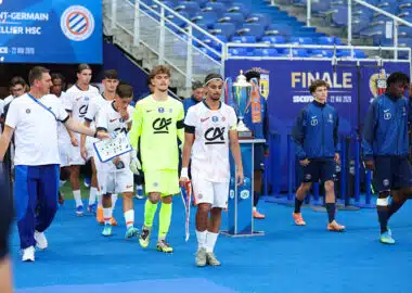 Soccer players in white uniforms walk on a blue field toward a trophy display during a final ceremony, with a neon-clad goalkeeper and staff nearby.