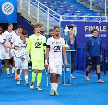 Soccer players in white uniforms walk on a blue field toward a trophy display during a final ceremony, with a neon-clad goalkeeper and staff nearby.