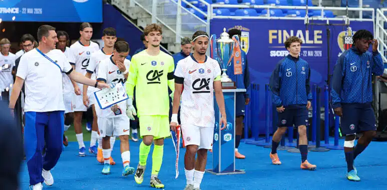 Gambardella | AllezPaillade.com Soccer players in white uniforms walk on a blue field toward a trophy display during a final ceremony, with a neon-clad goalkeeper and staff nearby.