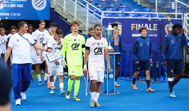 Soccer players in white uniforms walk on a blue field toward a trophy display during a final ceremony, with a neon-clad goalkeeper and staff nearby.
