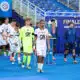 Soccer players in white uniforms walk on a blue field toward a trophy display during a final ceremony, with a neon-clad goalkeeper and staff nearby.