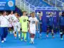 Soccer players in white uniforms walk on a blue field toward a trophy display during a final ceremony, with a neon-clad goalkeeper and staff nearby.