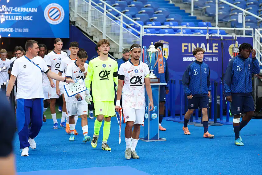 Gambardella | AllezPaillade.com Soccer players in white uniforms walk on a blue field toward a trophy display during a final ceremony, with a neon-clad goalkeeper and staff nearby.