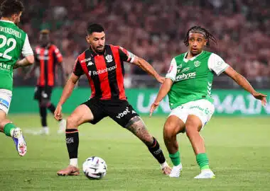 Two soccer players contesting the ball during a match; one wears red-and-black stripes, the other green-and-white kit, in a crowded stadium.